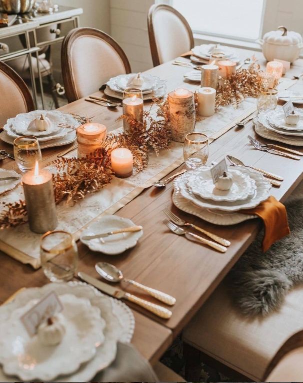 Styled Thanksgiving table featuring an elegant tablescape with white dinnerware, golden flatware, and warm candlelight. The table is adorned with a decorative runner, candles, and a centerpiece of greenery and small pumpkins.