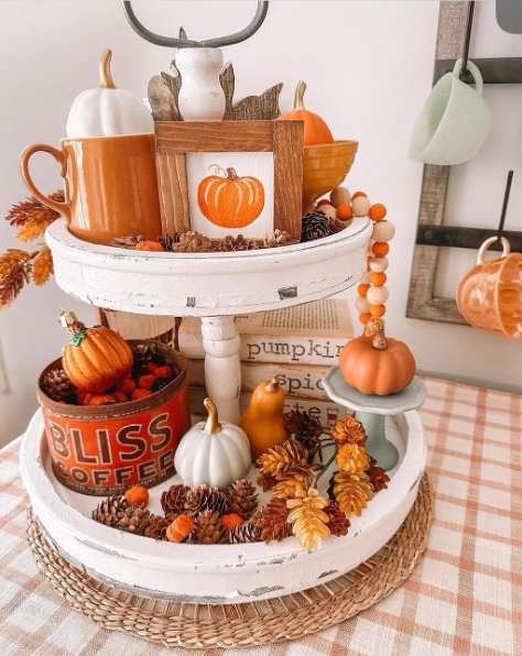A cozy tiered tray decorated for fall, featuring mini pumpkins, pinecones, and seasonal accents like an orange mug and a pumpkin-themed photo frame.