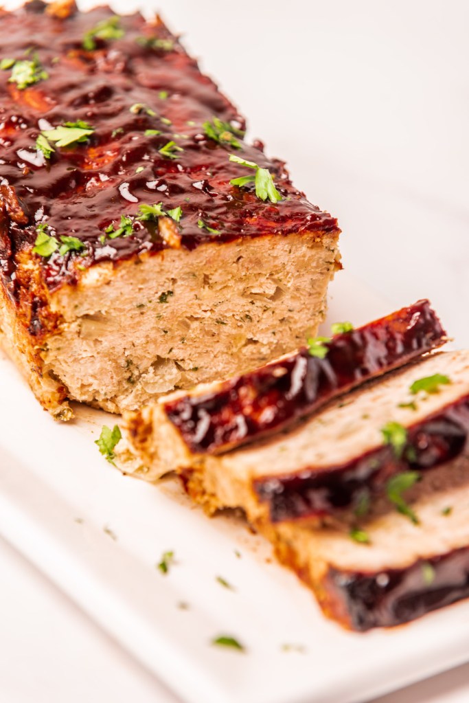 A close-up view of a sliced chicken meatloaf topped with a shiny glaze and garnished with chopped parsley, presented on a white plate.