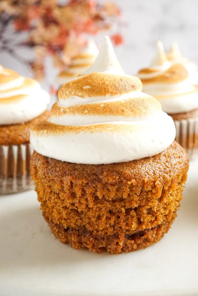 Close-up of a spiced pumpkin cupcake topped with a fluffy, toasted meringue frosting, set against a soft-focus autumnal background.