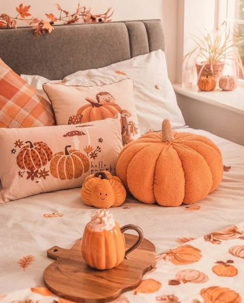 A cozy autumn-themed bedroom featuring a bed adorned with soft, pumpkin-patterned pillows and an oversized orange pumpkin plush. A wooden tray holds a pumpkin-shaped mug topped with whipped cream, along with small decorative pumpkins and scattered autumn leaves, creating a warm and inviting atmosphere.