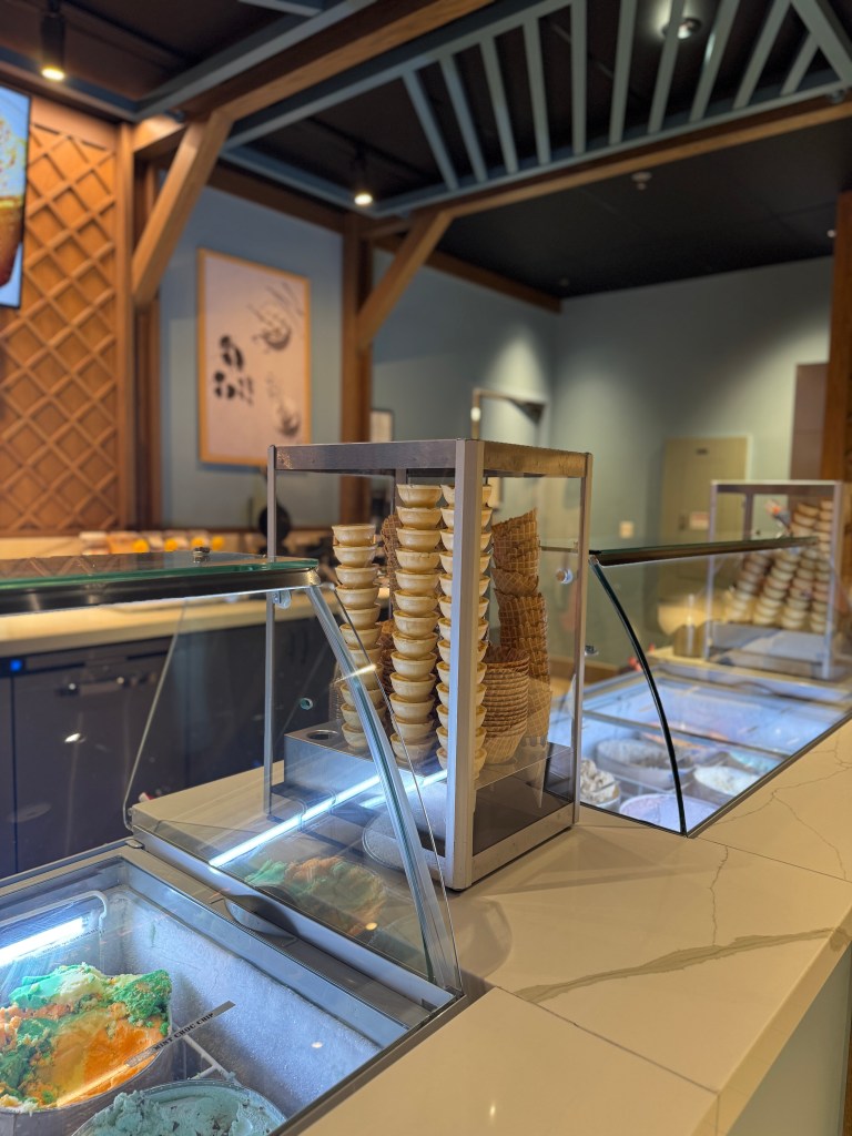 An ice cream shop display featuring colorful gelato flavors and stacked waffle cones, set in a cozy, modern environment.