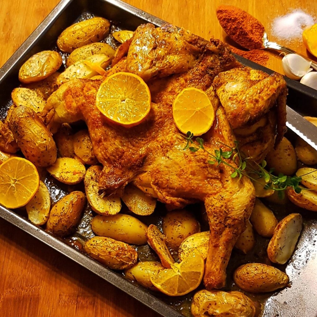 A roasted chicken arranged on a baking tray with golden potatoes and garnished with fresh lemon slices and herbs.