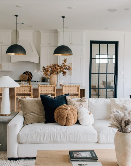 A cozy living room featuring a light-colored sofa adorned with various textured pillows, including a pumpkin-shaped cushion. In the background, a kitchen with pendant lights and a rustic dining area is visible, adding to the warm autumn atmosphere.