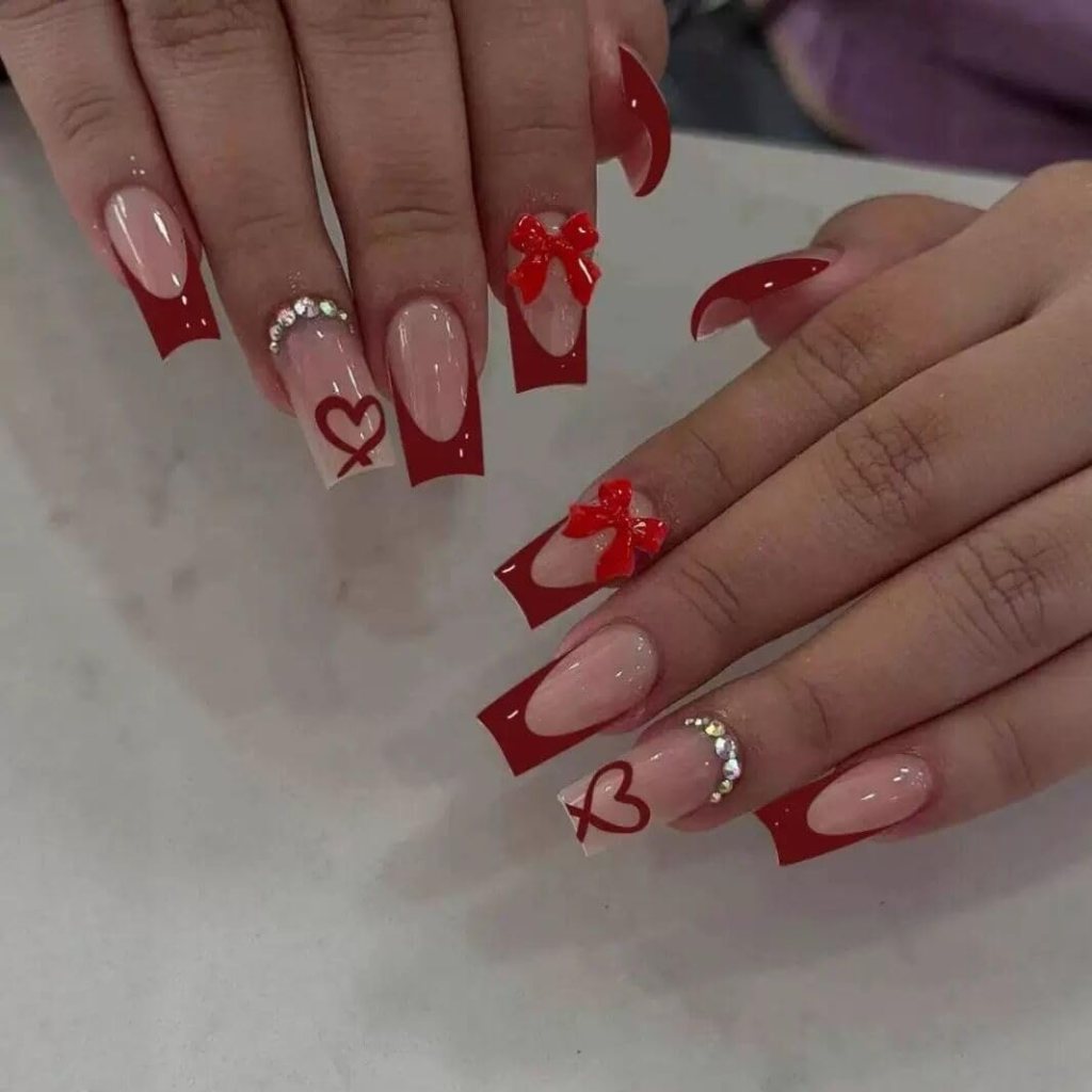 Close-up of hands with stylish red and nude long nails featuring heart and bow designs, along with rhinestone accents.
