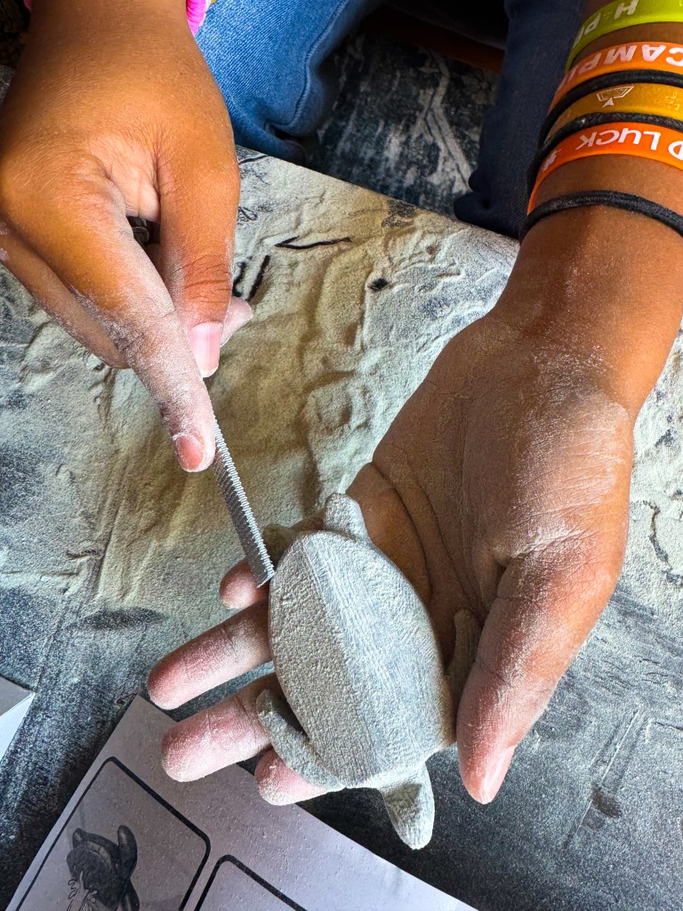 A person holding a small, unfinished turtle sculpture in one hand while using a tool to carve it. The hand is covered in fine dust from the carving process, and there are colorful wristbands visible on the other wrist.