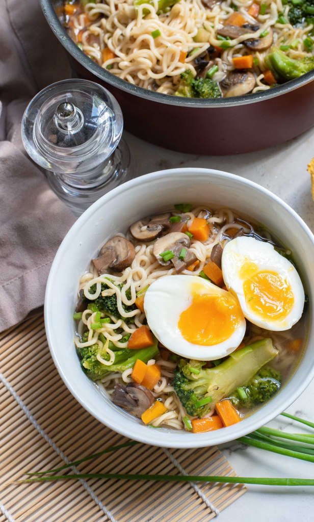 A bowl of ramen noodles topped with sliced soft-boiled eggs, broccoli, carrots, and mushrooms, served alongside a pot of simmering soup.