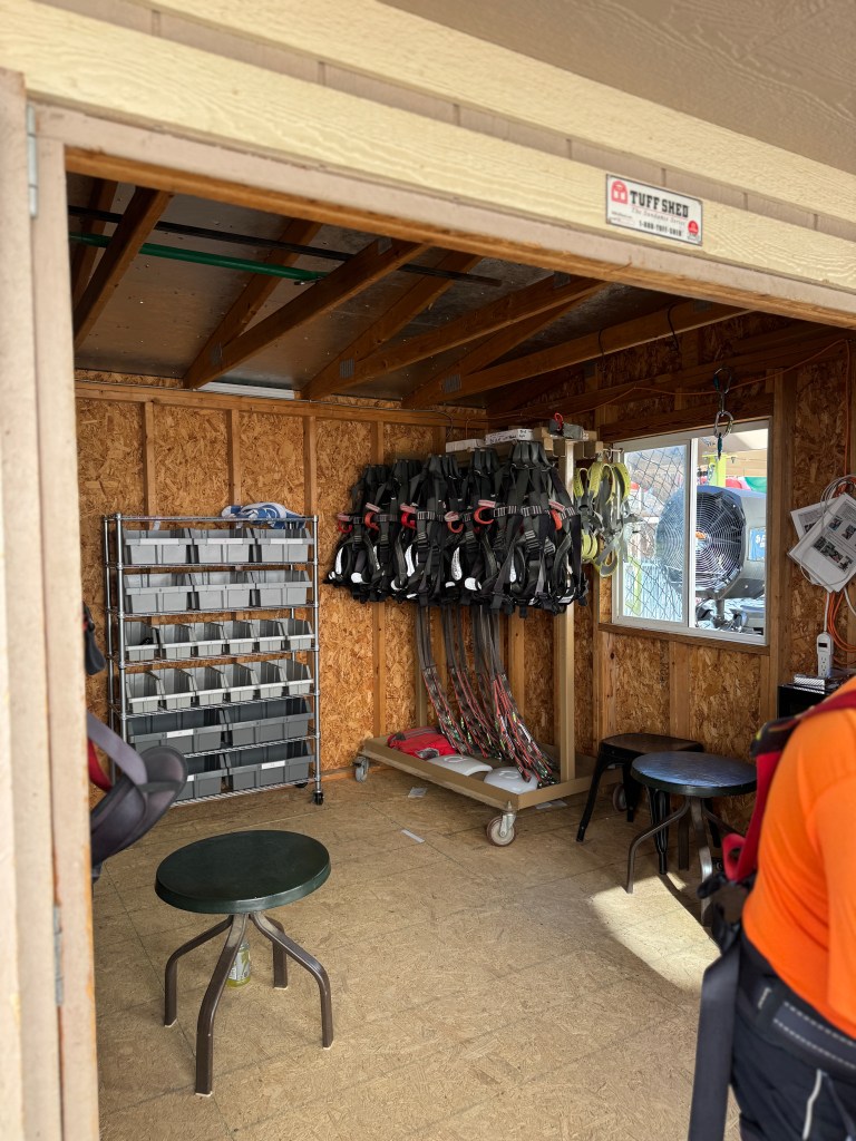 An indoor facility at Great Wolf Lodge featuring harnesses hanging on a rack, shelving with storage boxes, and seating areas. The room has wooden walls and an open view towards the outdoor adventure area.