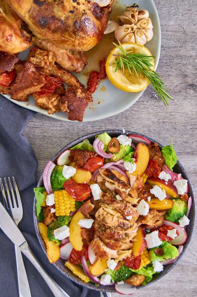 A roasted chicken served on a plate alongside garlic, lemon, and herbs, with a colorful salad featuring grilled chicken, mixed greens, corn, peaches, tomatoes, and feta cheese in a bowl.