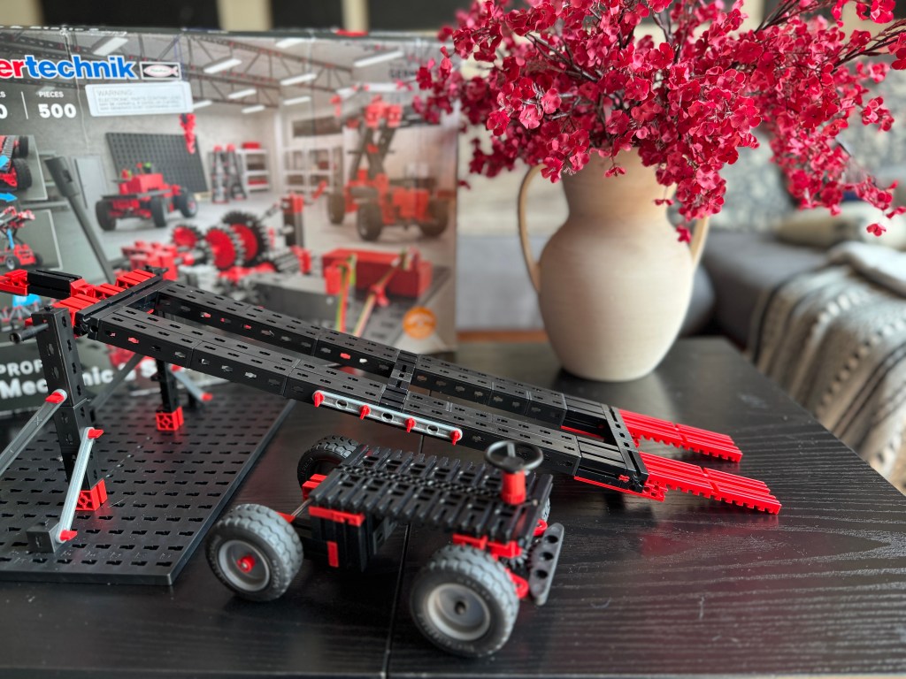 An assembled Fischer Technik STEM kit featuring a vehicle and ramp on a dark table, with a background of floral decor and a product box.