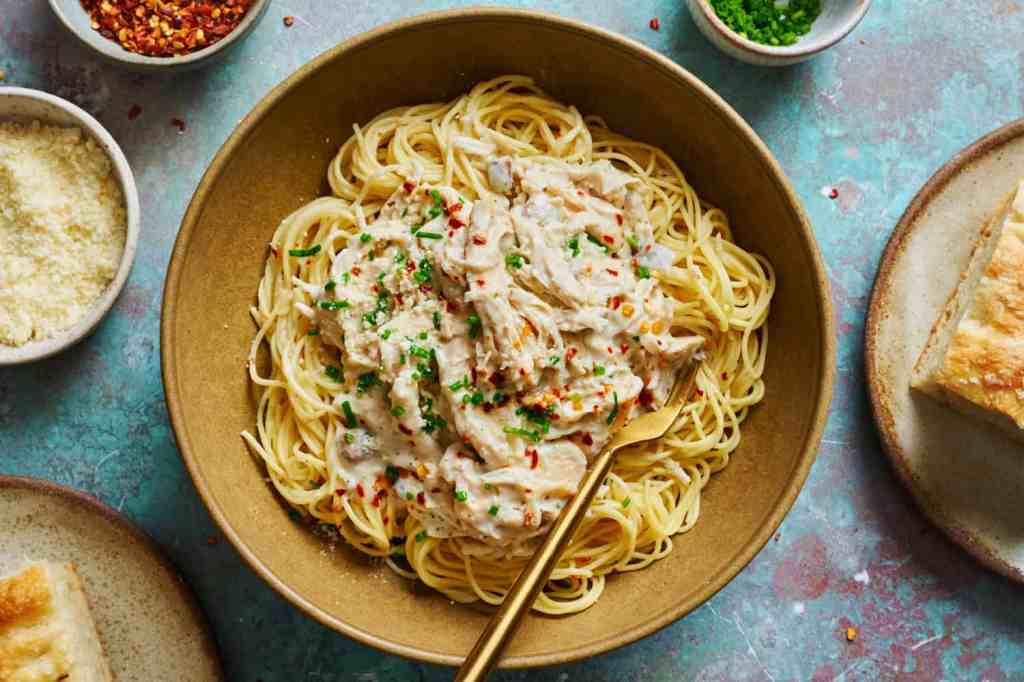 A bowl of pasta topped with creamy chicken, garnished with chopped herbs and red pepper flakes, accompanied by slices of bread on the side.