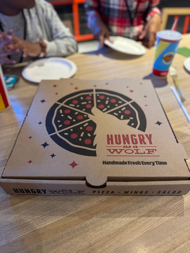 A pizza box from Hungry as a Wolf, featuring a graphic of a wolf and pizza design, placed on a wooden table alongside plates and drinks.