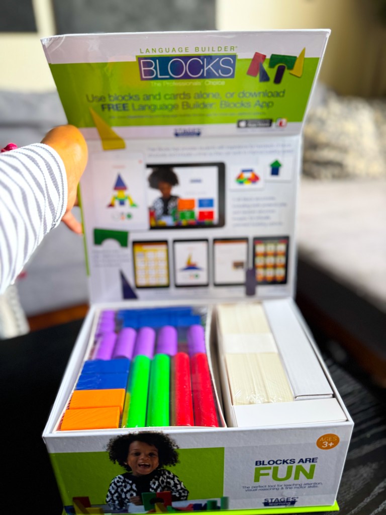 A colorful educational box of Language Builder Blocks, featuring various blocks and cards, accompanied by an advertisement for the Language Builder Blocks app. The box is being held by a person, with a child smiling in the foreground.