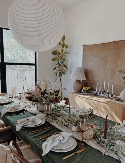 A cozy fall dining table setup featuring a green tablecloth, white dishes, and a centerpiece of greenery and candles, complemented by wooden accents and a paper lantern above.