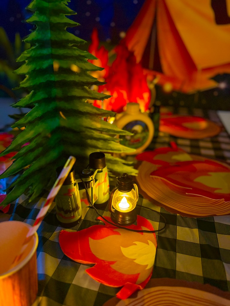 Moody camping party table setup with lanterns, fire plates, and woodland decorations.