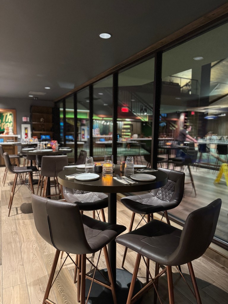 Interior view of a restaurant featuring sleek tables with dark chairs, set for dining, illuminated by soft lighting.