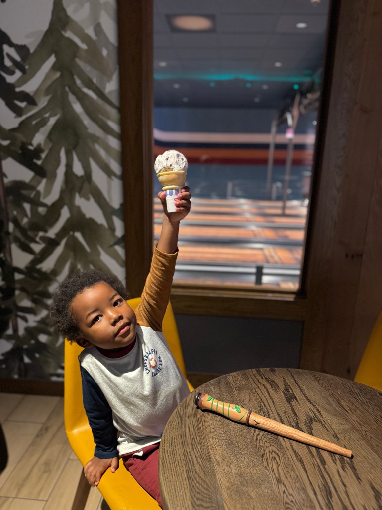 A young child sitting at a wooden table inside a cozy restaurant, happily holding up an ice cream cone with a cheerful expression. Next to them is a whimsical wooden toy resembling a stick with decorative features.
