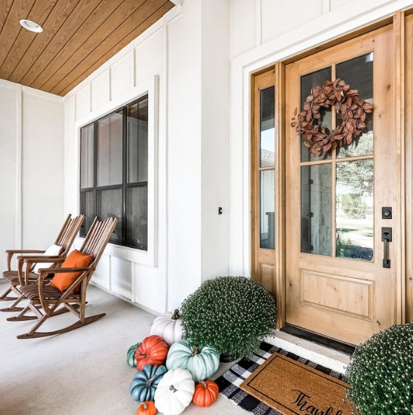 A cozy porch featuring two wooden rocking chairs with orange cushions, a welcoming front door adorned with a floral wreath, and colorful pumpkins arranged on the steps beside a green plant.