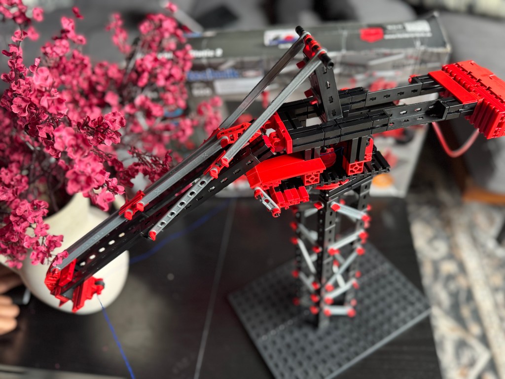 A close-up view of a Fischer Technik STEM kit model, featuring a crane-like structure in black and red, surrounded by blooming pink flowers and a box in the background.