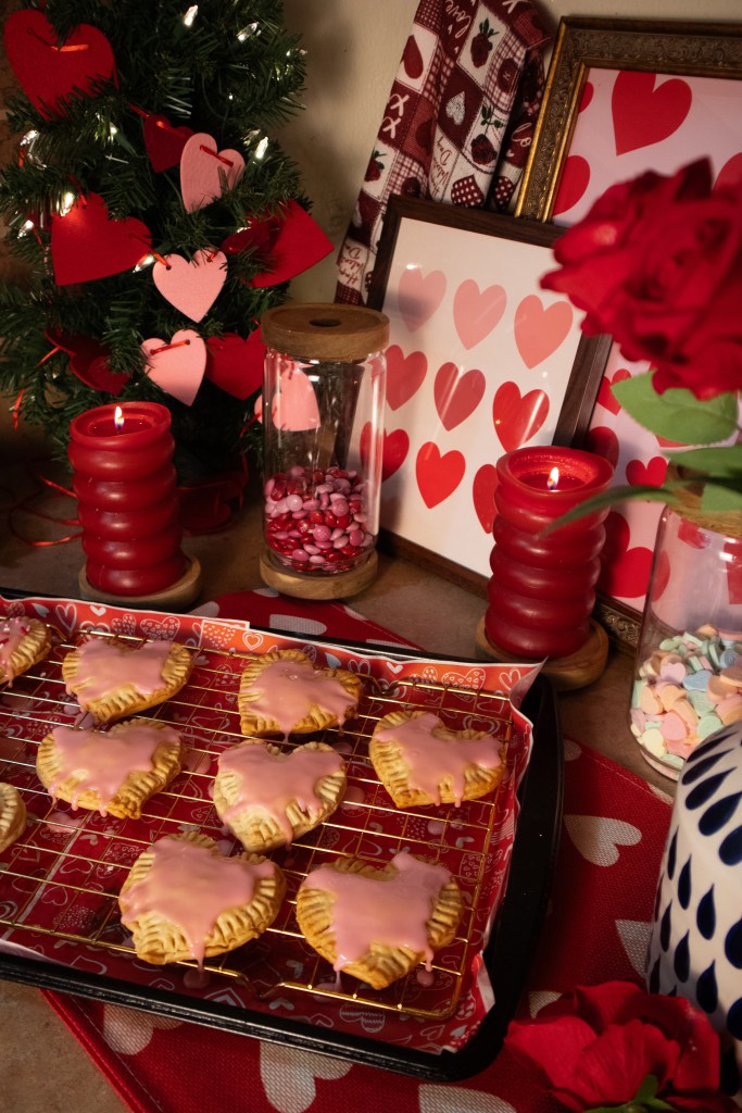 A cozy Valentine's Day display featuring heart-shaped pop tarts with pink icing, red candles, a small decorated tree with heart ornaments, and decorative jars filled with colorful candies. In the background, framed images with heart patterns and a bouquet of red roses complete the festive atmosphere.
