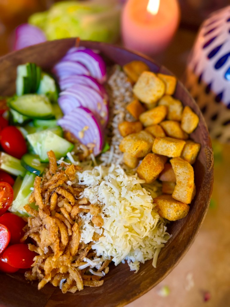 A wooden bowl filled with a colorful salad featuring sliced cucumbers, cherry tomatoes, red onions, crispy fried onions, shredded cheese, croutons, and grains, with a flickering candle in the background.