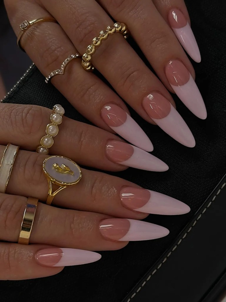 Close-up of hands with long, pointed pink and nude ombre nails adorned with multiple rings, including gold and pearl details.