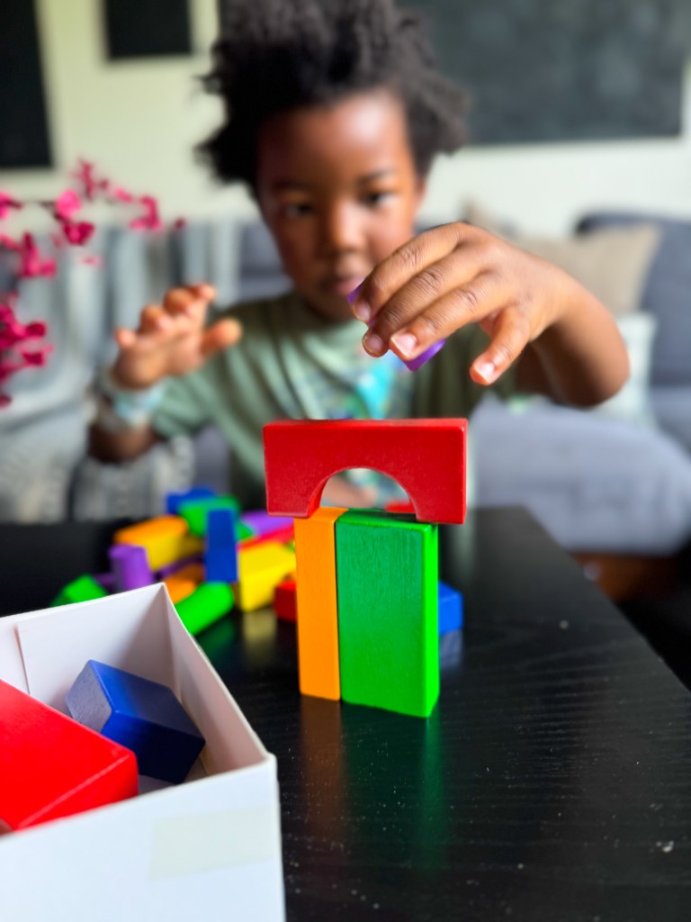 A child building a colorful structure with wooden blocks on a black table, with a box of assorted blocks visible in the foreground.