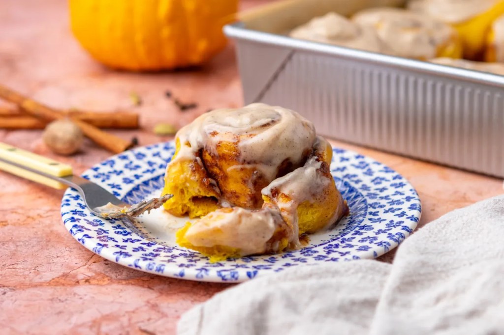 Close-up of a frosted pumpkin cinnamon roll on a patterned plate, with a fork beside it, on a table adorned with cinnamon sticks and a small pumpkin in the background.