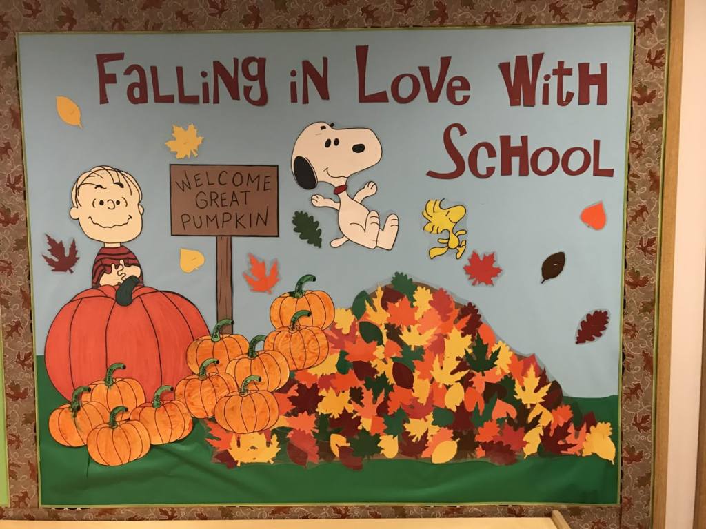 Falling in Love With School bulletin board featuring pumpkins, a large pile of autumn leaves and playful fall classroom decorations.