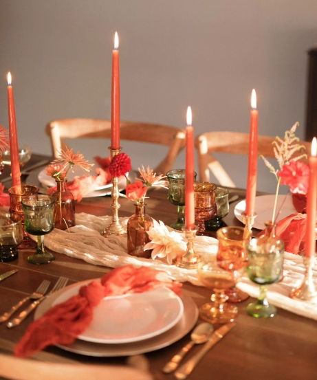 A cozy fall tablescape featuring warm orange candles, colorful glassware, and floral accents. The setting includes a simple white table runner and adorned plates, creating an inviting atmosphere.
