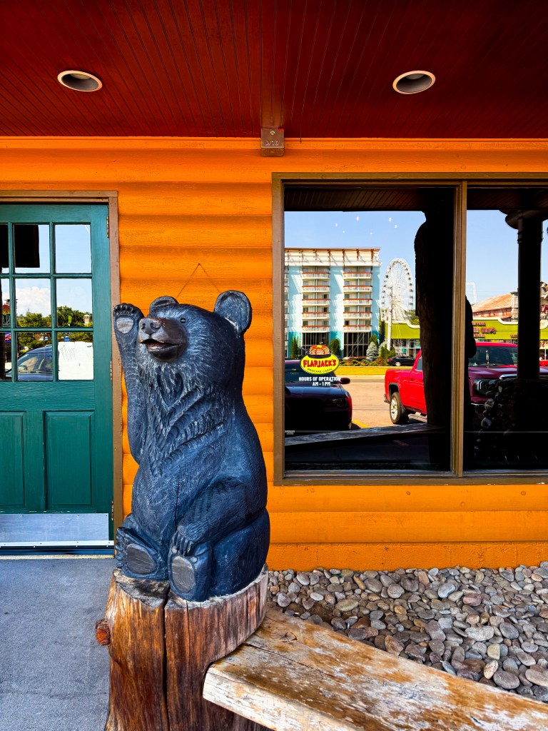 Black bear statue outside Flapjack's Pancake Cabin with orange log cabin siding and a green entrance door.