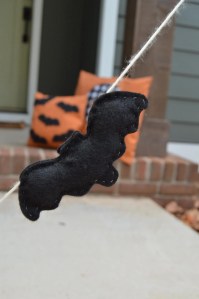 A black felt bat hanging from a string, with colorful Halloween pillows in the background.