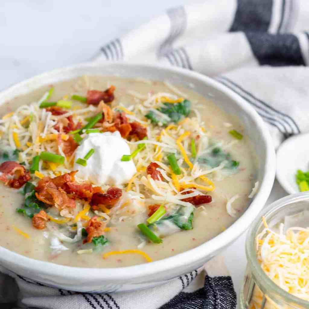 A bowl of creamy potato soup topped with shredded cheese, crumbled bacon, chopped green onions, and a dollop of sour cream, served alongside a striped cloth.