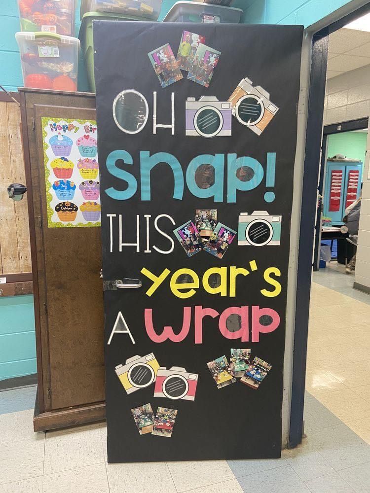 A classroom door decorated with a black poster displaying the phrase 'OH SNAP! THIS YEAR'S A WRAP' in colorful letters, accompanied by camera illustrations and several photos attached to the poster.