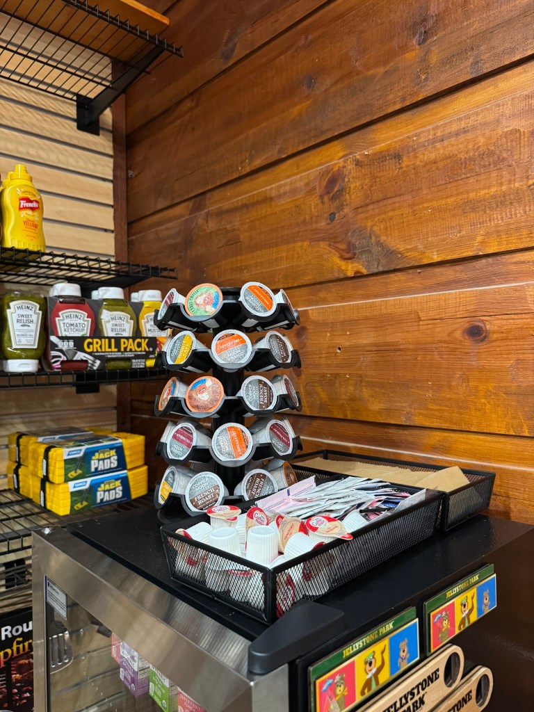 A display of condiment cups stacked on a black tray, surrounded by various sauces and condiments on metal shelves against a wooden wall.