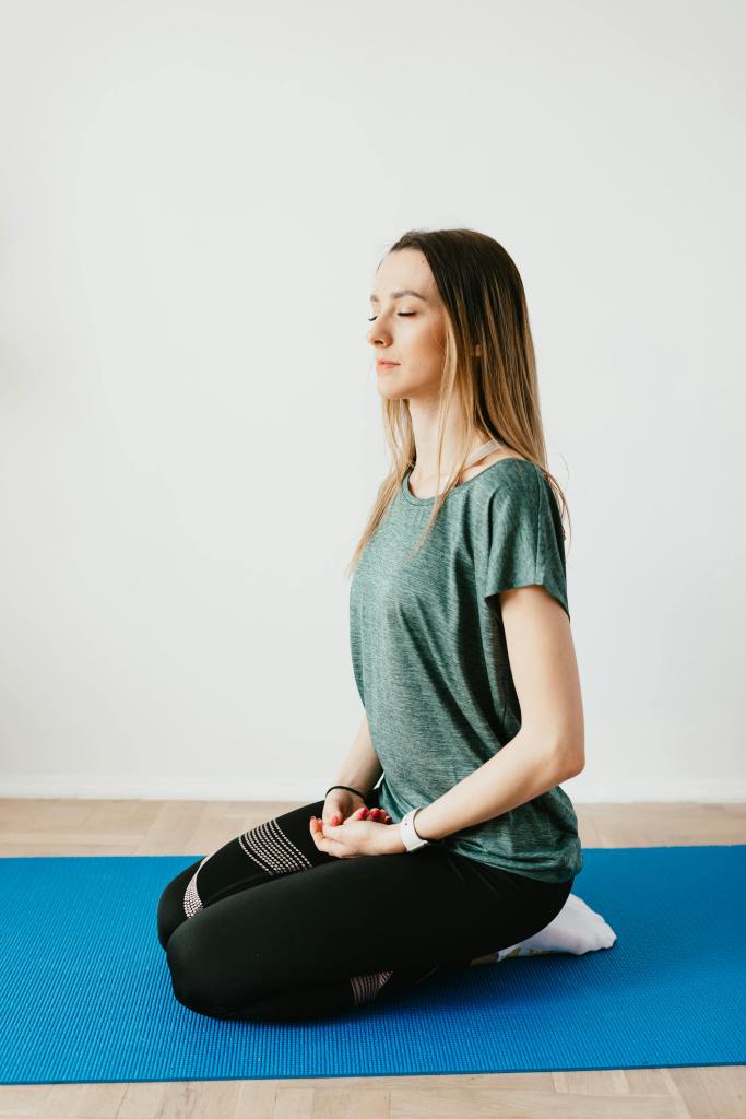 A woman meditating on a blue yoga mat, eyes closed in a relaxed position, wearing comfortable athletic clothing.