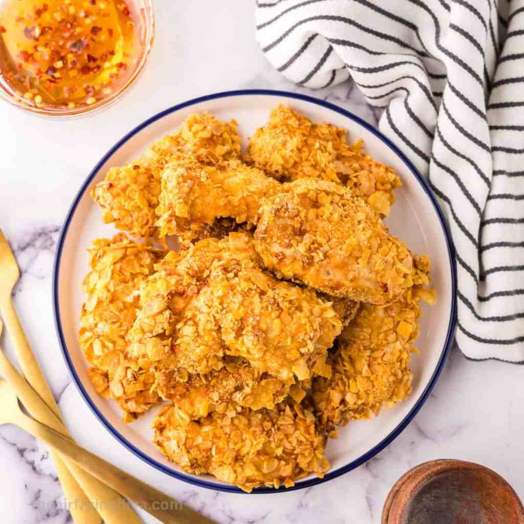 A plate of crispy fried chicken pieces coated in cornflakes, served with a bowl of dipping sauce and a striped napkin in the background.