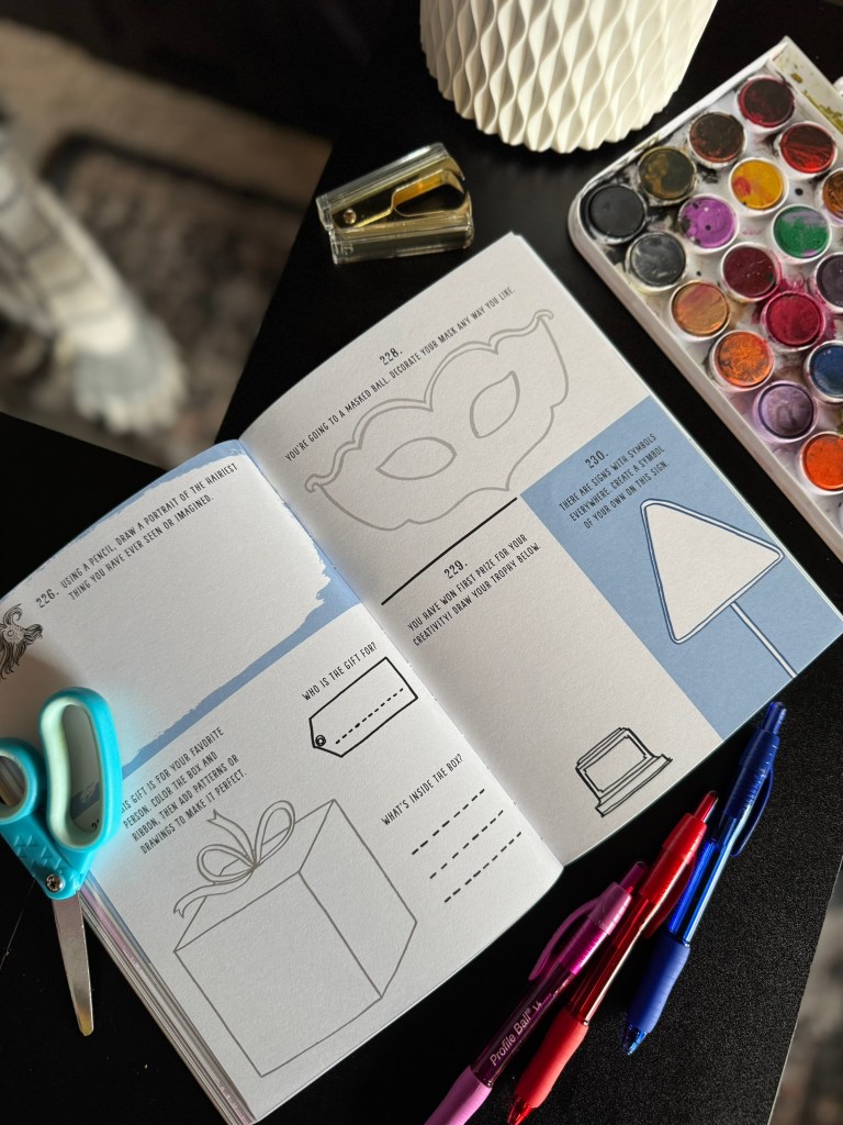An open art book with prompts on its pages, featuring illustrations of a mask, a present, and a lamp. Nearby are scissors, colorful pens, and a watercolor palette.