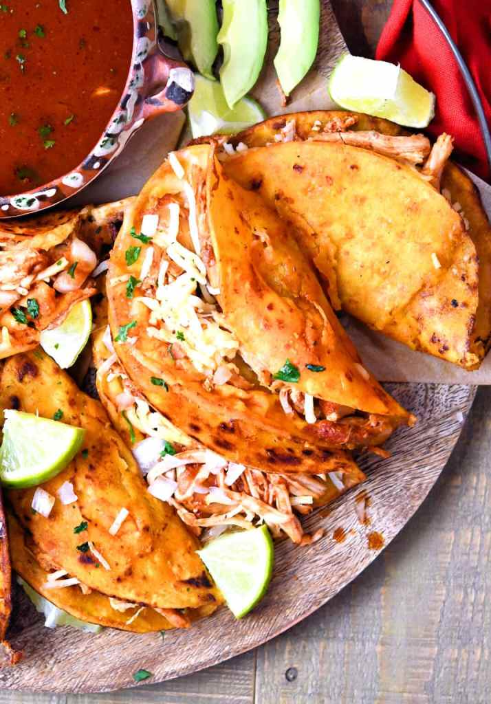 A platter of chicken birria tacos with shredded chicken inside crispy tortillas, garnished with cheese and fresh cilantro, accompanied by lime wedges and a bowl of dipping sauce.