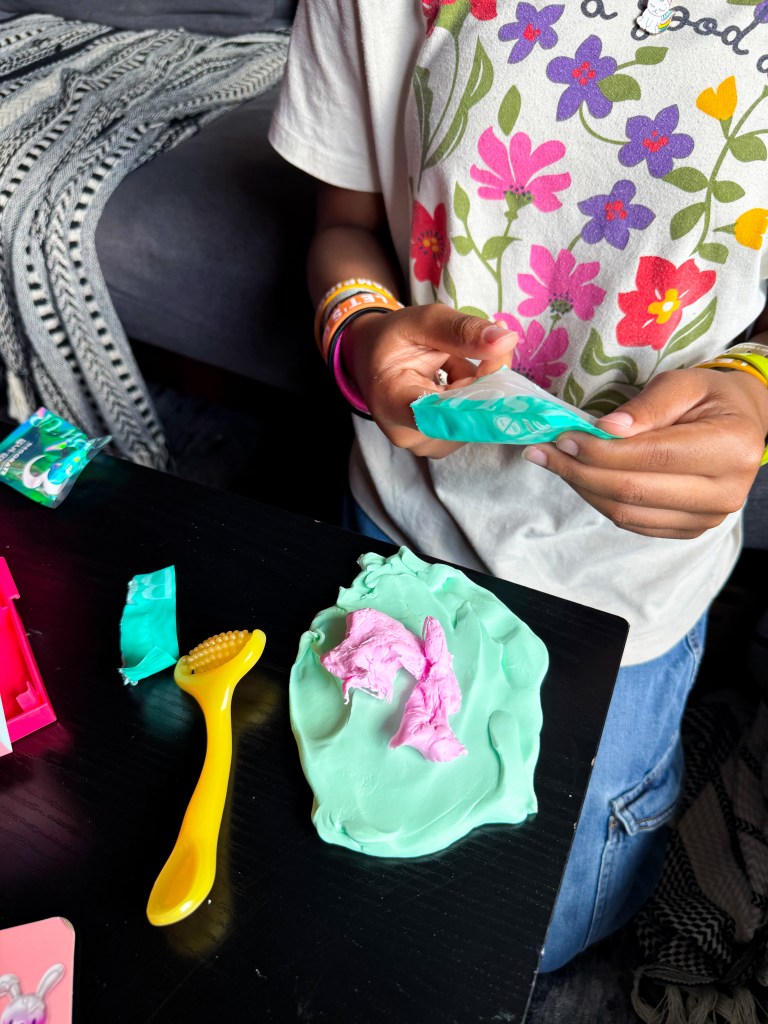 A child holds a package of colorful modeling clay while playing with mint-green clay on a black table, accompanied by a yellow shaping tool and colorful wrappers.