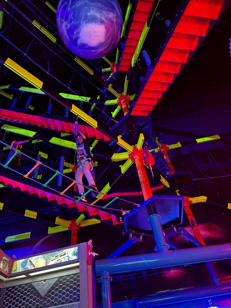 An indoor adventure park featuring colorful climbing structures and a person navigating a high rope course, with a planet-like decoration overhead.