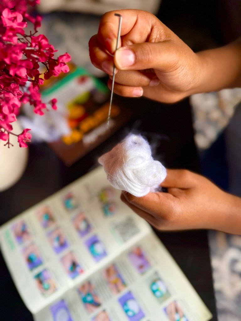 A close-up of a person's hand holding a felting needle over a ball of wool, with a colorful instruction booklet and pink flowers in the background.