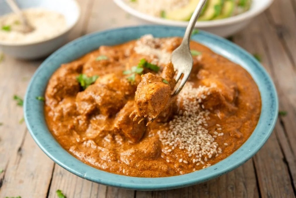 A bowl of chicken curry garnished with sesame seeds and fresh herbs, with a fork lifting a piece of chicken above the dish.