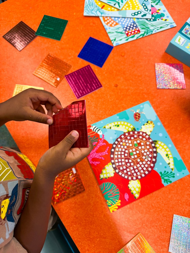 Child peeling a red mosaic sticker sheet while completing a hands-on kindergarten project.