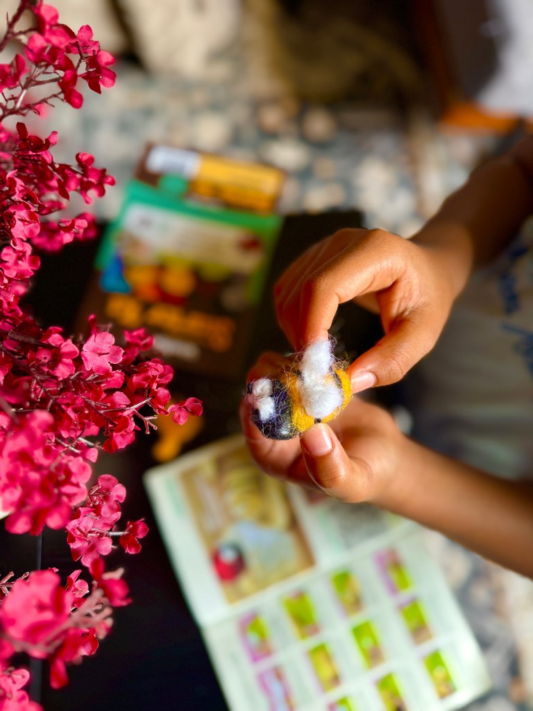 A close-up image of a child's hands holding a colorful felted bee made from wool, with a craft instruction booklet and artificial pink flowers in the background.