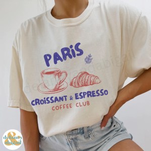 A person wearing a white t-shirt featuring a graphic design that reads 'PARIS Croissant & Espresso Coffee Club' with illustrations of a coffee cup and a croissant, paired with denim shorts.
