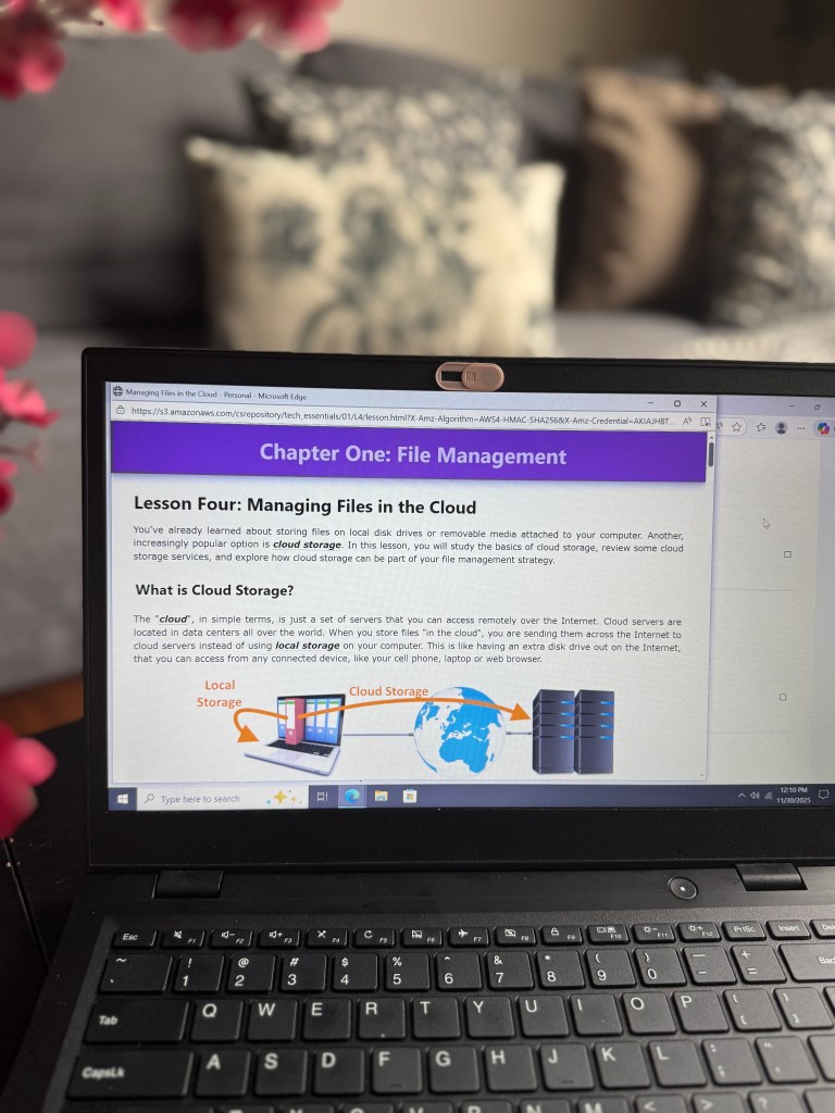 A laptop screen displaying a lesson on 'Managing Files in the Cloud' from a tech curriculum, with purple headings and content explaining cloud storage and its importance, set against a background of a living room.