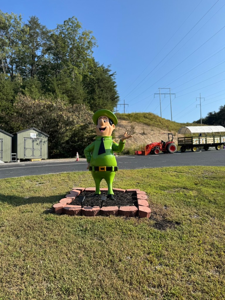 A cheerful green cartoon character with a hat and a smile, standing on a planted area surrounded by brick borders, against a backdrop of trees and a clear blue sky.