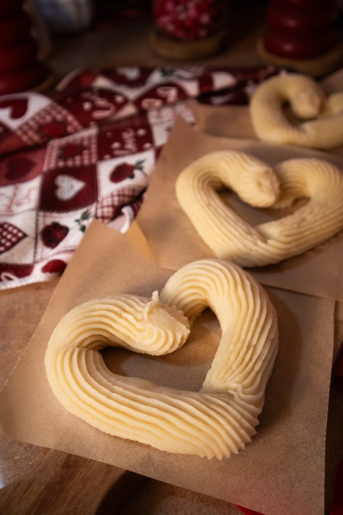 Three shaped pieces of buttercream frosting arranged in heart shapes, placed on brown parchment paper, with a patterned red and white cloth in the background.