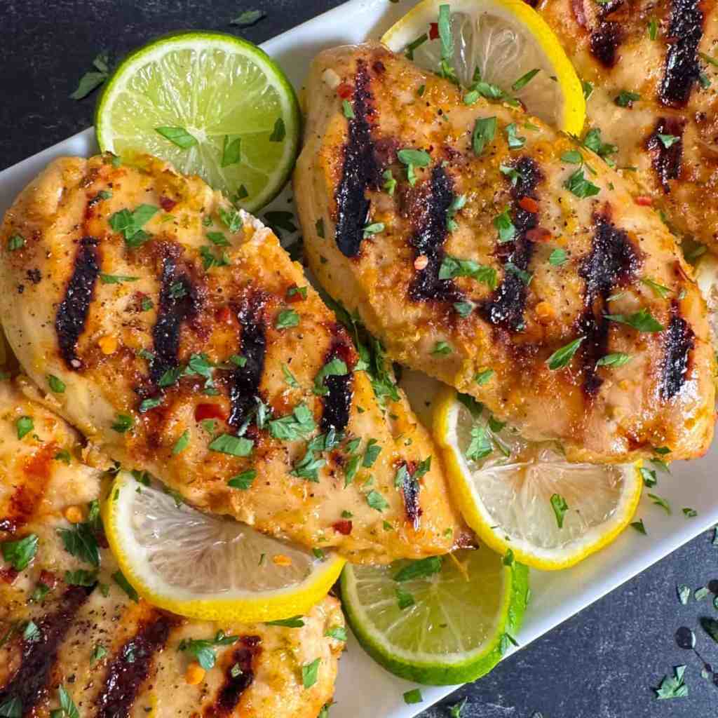 Grilled chicken breasts garnished with fresh parsley, served with lemon and lime slices.